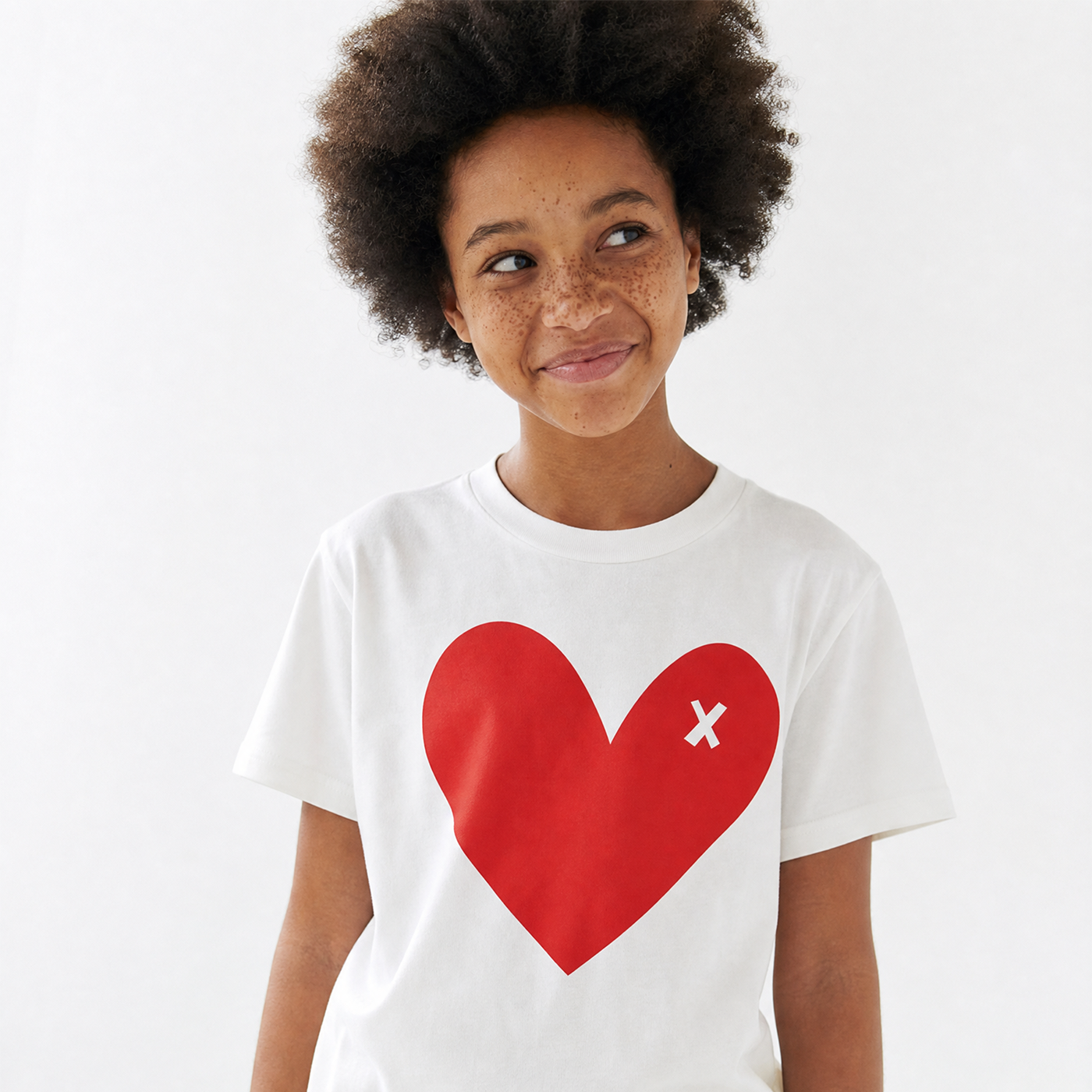 A young girl with dark curly hair and freckles wearing a white short-sleeve t-shirt with a large red heart graphic on the front featuring a small white 'x' inside the heart, set against a plain white background.