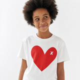 A young girl with dark curly hair and freckles wearing a white short-sleeve t-shirt with a large red heart graphic on the front featuring a small white 'x' inside the heart, set against a plain white background.