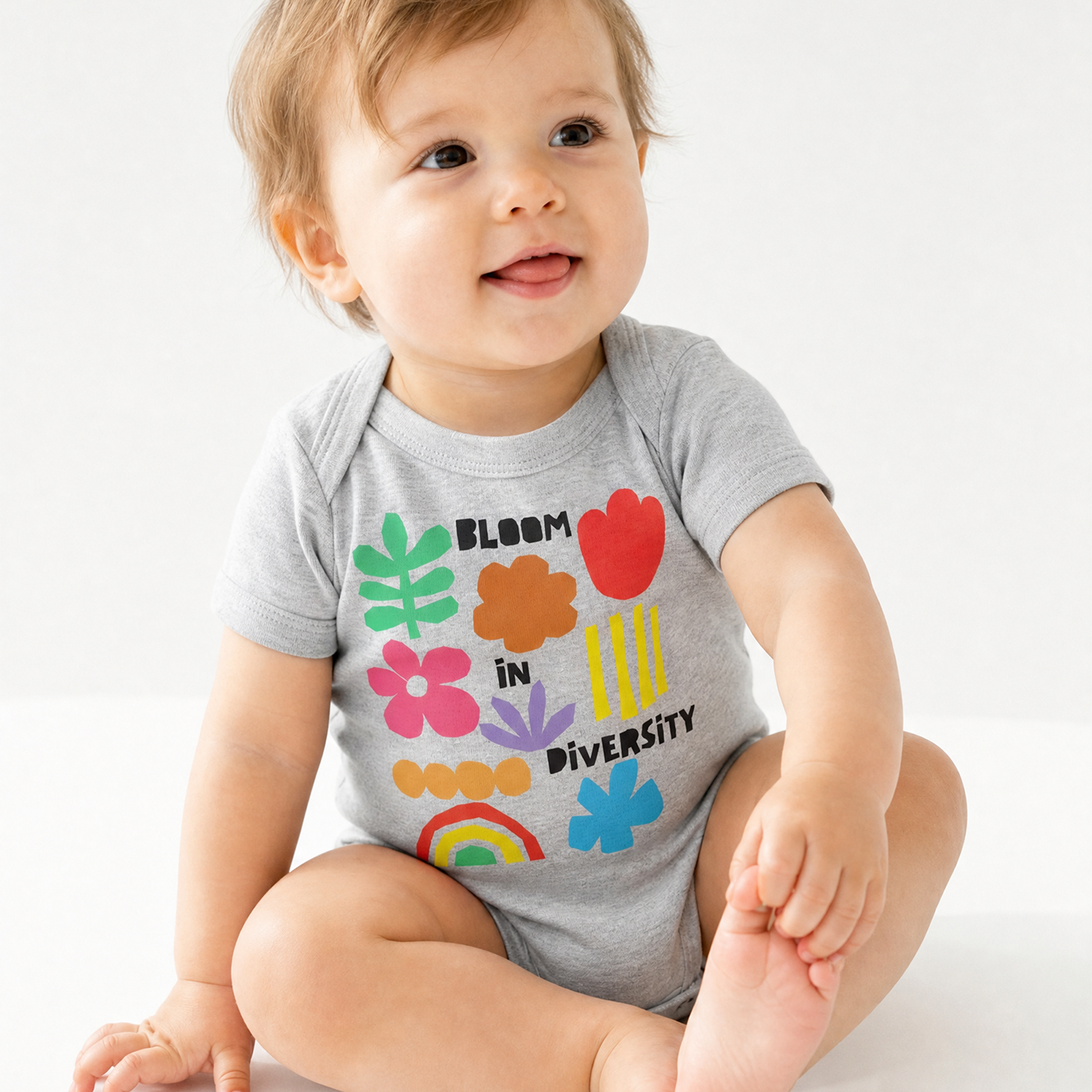 Smiling baby sitting in a heather gray "Bloom in Diversity" bodysuit featuring colorful flower, leaf, and rainbow graphics, against a white background.
