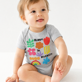 Smiling baby sitting in a heather gray "Bloom in Diversity" bodysuit featuring colorful flower, leaf, and rainbow graphics, against a white background.