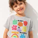 Smiling toddler girl wearing a heather gray "Bloom in Diversity" tee with colorful flower, leaf, and rainbow graphics, against a sunlit white wall.