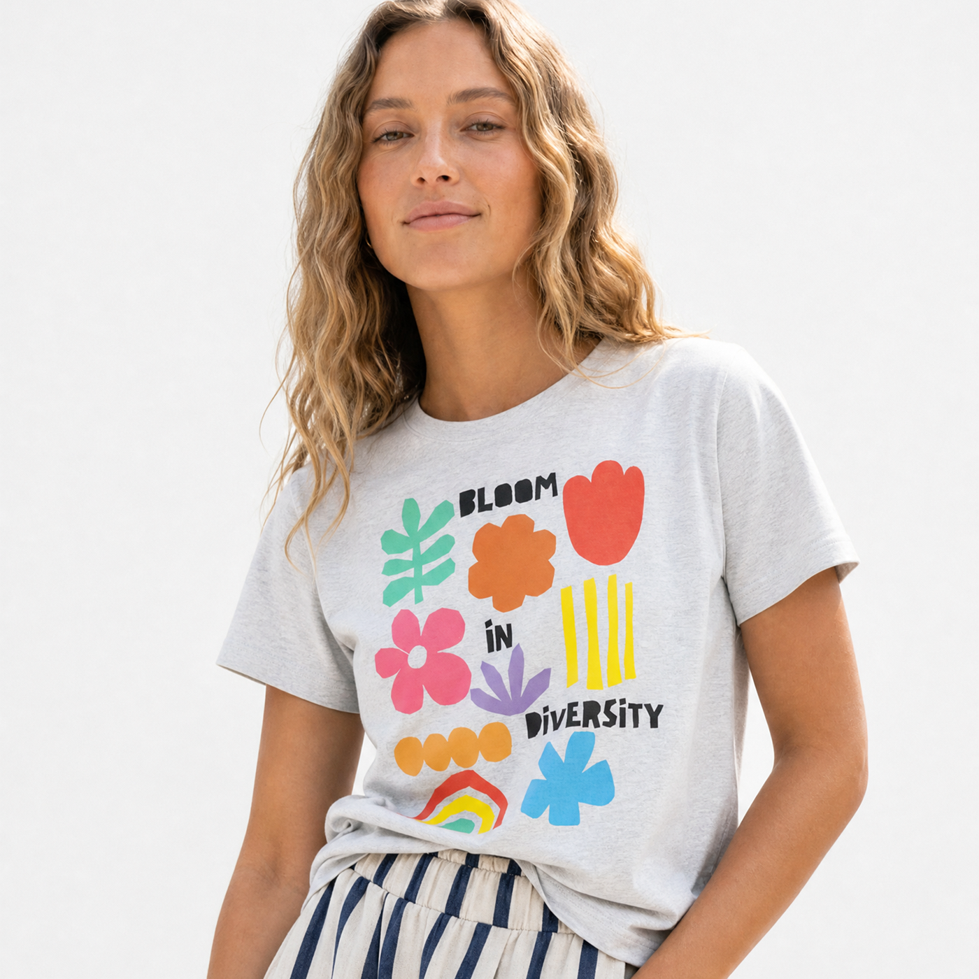 Woman with wavy blonde hair wearing a heather gray "Bloom in Diversity" tee with colorful floral graphics, paired with navy-and-cream striped pants.