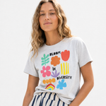 Woman with wavy blonde hair wearing a heather gray "Bloom in Diversity" tee with colorful floral graphics, paired with navy-and-cream striped pants.