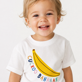 Smiling toddler with blond curly hair wearing a white "Cuckoo Bananas" tee featuring a big yellow banana graphic and colorful lettering.