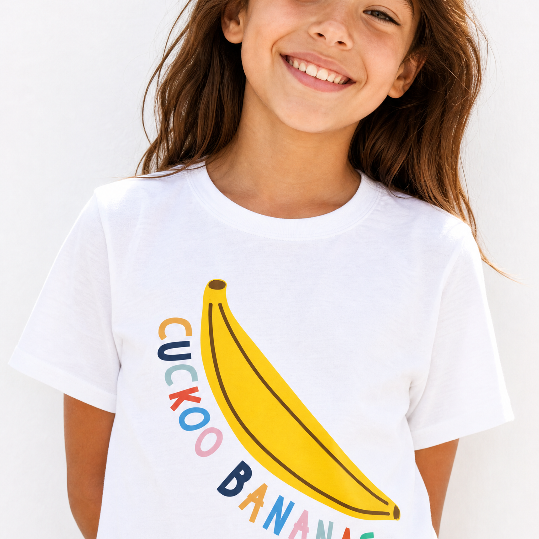 A young girl smiling while wearing a white t-shirt with a graphic of a yellow banana and the text 'CUCKOO BANANAS' in colorful letters.