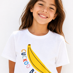A young girl with long brown hair smiling while wearing a white crewneck t-shirt with a yellow banana graphic and the text 'CUCKOO BABY' in colorful letters.