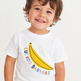 A smiling toddler wearing a white short-sleeve t-shirt with a yellow banana graphic and the words 'CUCKOO BANANAS' in colorful letters, paired with light-colored patterned shorts against a plain white background.
