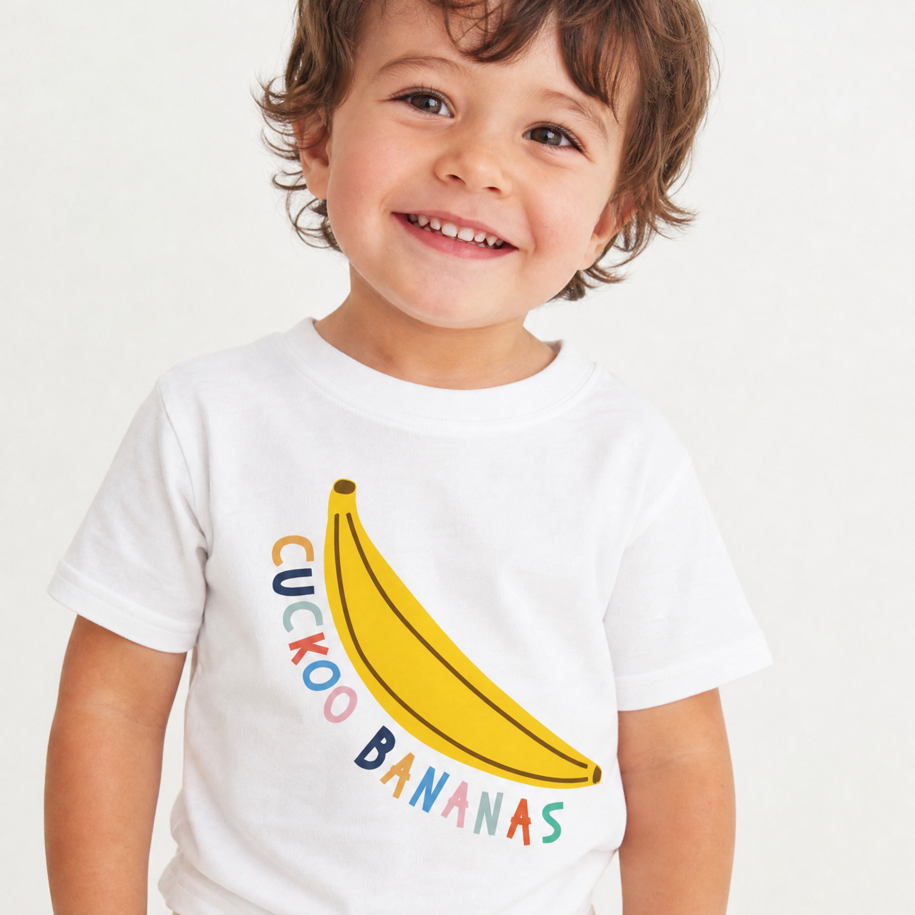A smiling toddler wearing a white short-sleeve t-shirt with a yellow banana graphic and the words 'CUCKOO BANANAS' in colorful letters, paired with light-colored patterned shorts against a plain white background.