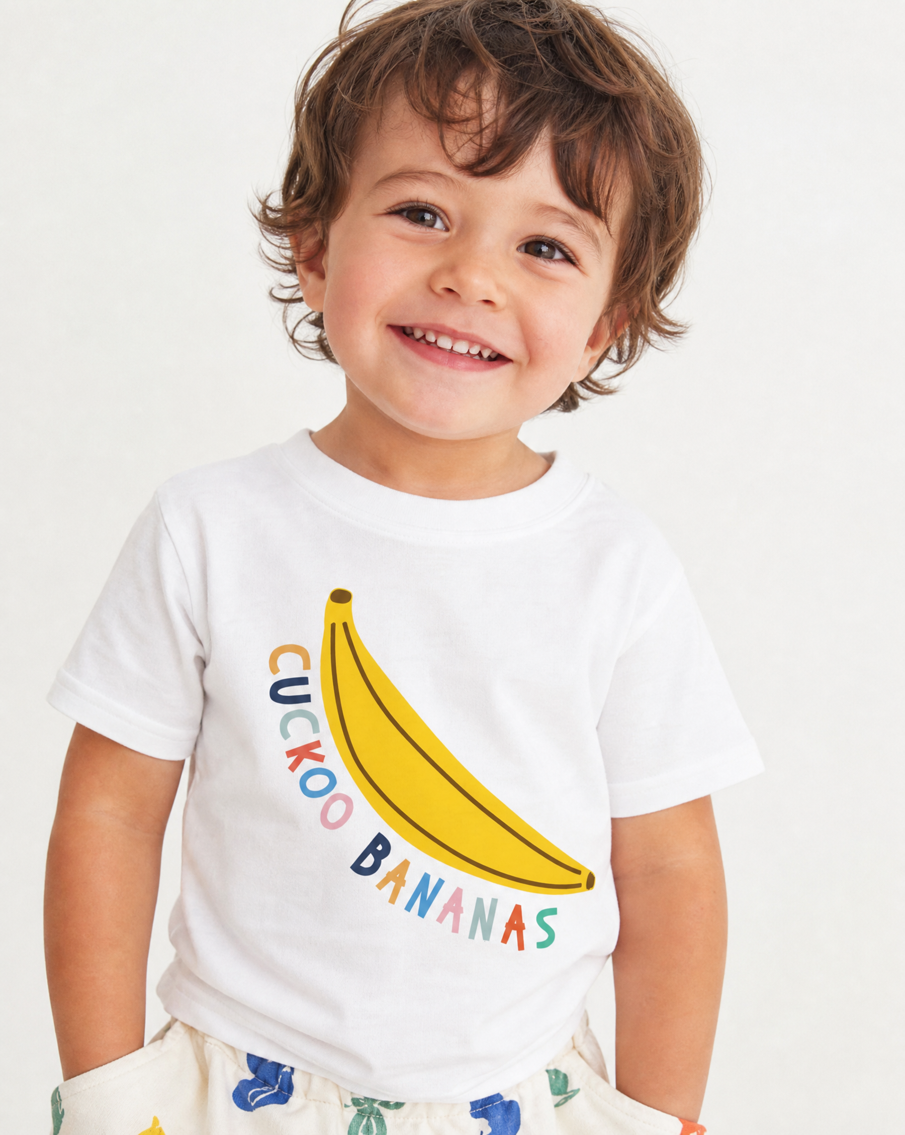A smiling toddler wearing a white short-sleeve t-shirt with a yellow banana graphic and the words 'CUCKOO BANANAS' in colorful letters, paired with light-colored patterned shorts against a plain white background.