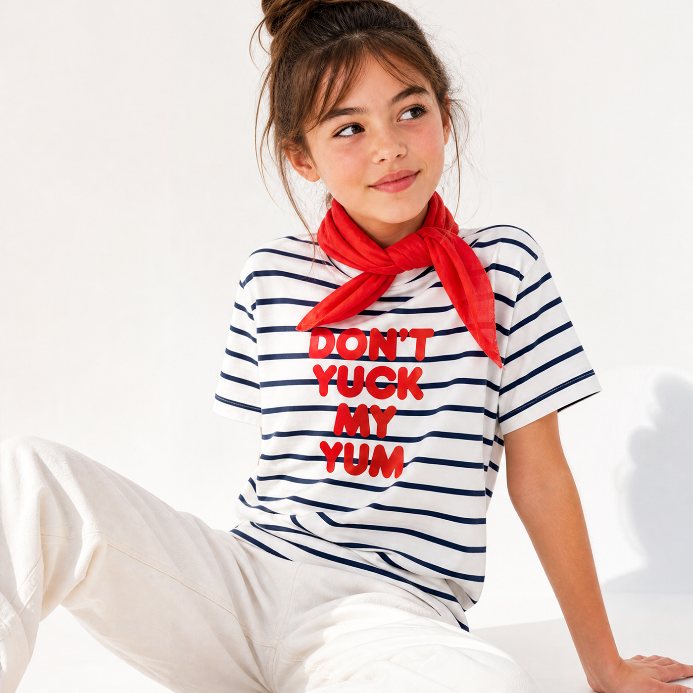 A young girl wearing a white short-sleeve t-shirt with navy blue horizontal stripes and bold red text that reads 'DON'T YUCK MY YUM'. She is also wearing a bright red neck scarf and white pants, sitting against a clean white background.