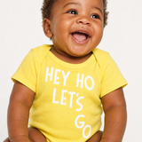Laughing baby wearing a yellow "Hey Ho Let's Go" bodysuit with white lettering, against a light background.