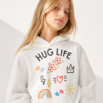 A blonde woman wearing a white pullover hoodie with a colorful graphic print that reads 'HUG LIFE' above flowers, stars, a crown, lightning bolts, a heart, a rainbow, and a sun, photographed against a clean white background.