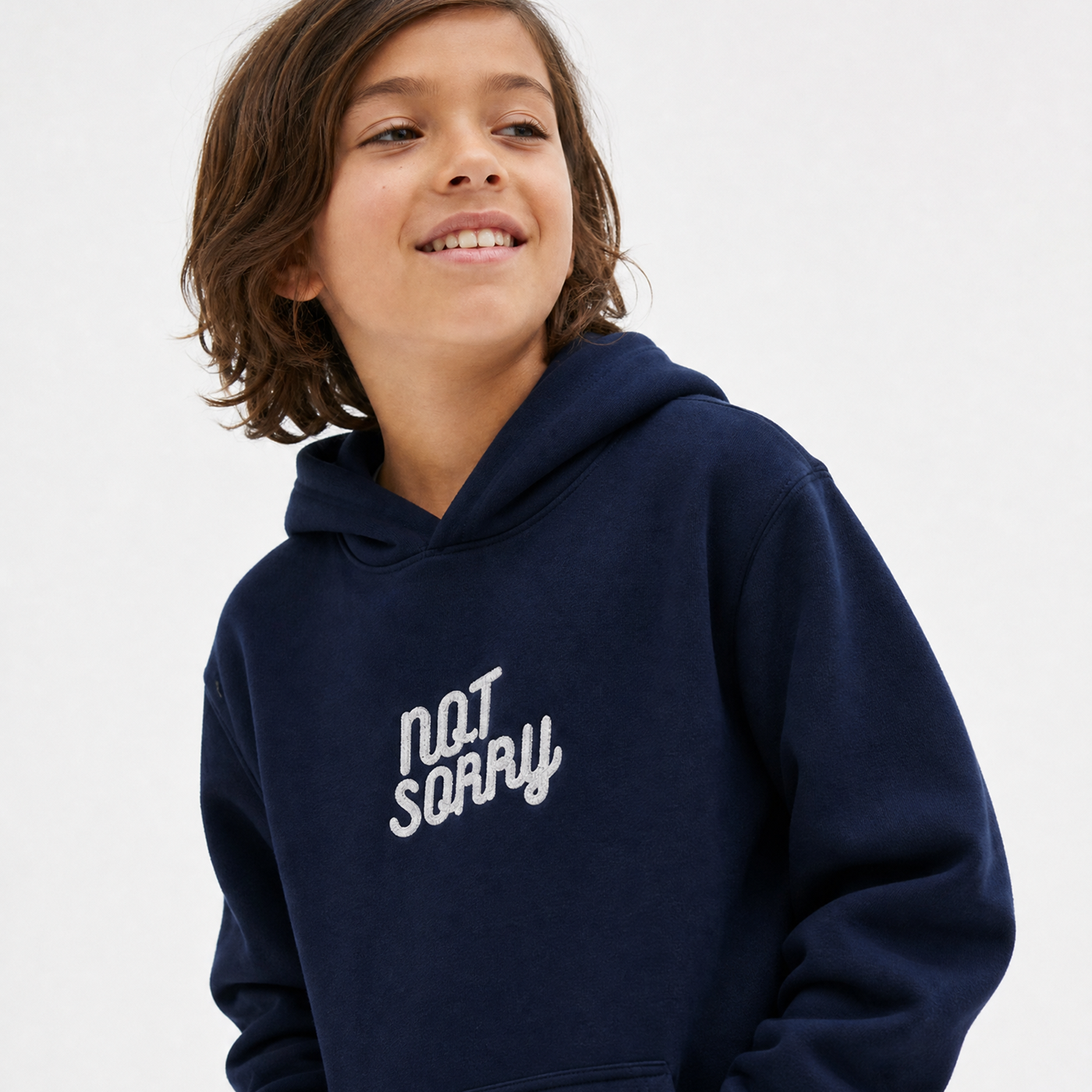 A smiling young boy with brown hair wearing a navy blue hoodie with 'NOT SORRY' embroidered in white script on the chest, set against a plain light gray background.