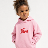 A young child with curly hair wearing a light pink hoodie with the words 'NOT SORRY' embroidered in red across the chest, set against a plain white background.