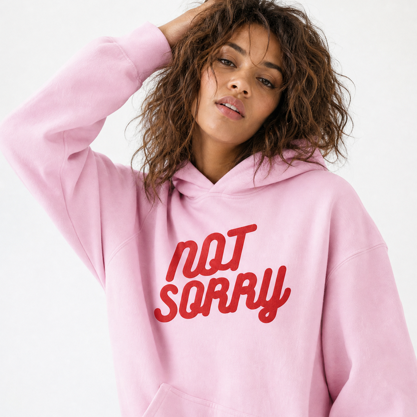 A woman wearing a light pink hoodie with the words 'NOT SORRY' printed in red across the chest, posed against a plain white background.