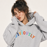 A woman wearing a light gray oversized hoodie with the word 'OBVIOUSLY' printed across the chest in colorful letters, posing against a plain off-white background.
