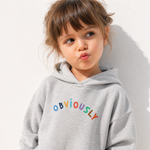 A young girl with brown hair wearing a light gray heathered hoodie with the word 'OBVIOUSLY' printed across the chest in colorful letters, posing against a white wall.