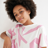 A close-up of a person with dark curly hair wearing a white short-sleeve T-shirt with a pink abstract brushstroke pattern, photographed against a plain light background.