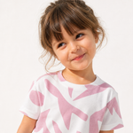 A young girl with brown hair and bangs smiling while wearing a white short-sleeve t-shirt with a pink abstract pattern, set against a plain white background.