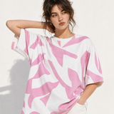 A woman wearing a loose white short-sleeve T-shirt with a bold pink abstract brushstroke pattern, posed against a bright white wall.
