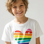 Smiling boy with tousled blond hair wearing a white T-shirt with a large rainbow-striped heart graphic, against a light gray background.