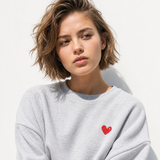 A woman with short brown hair wearing a light gray crewneck sweatshirt with a small red heart on the left chest, photographed against a plain white background.