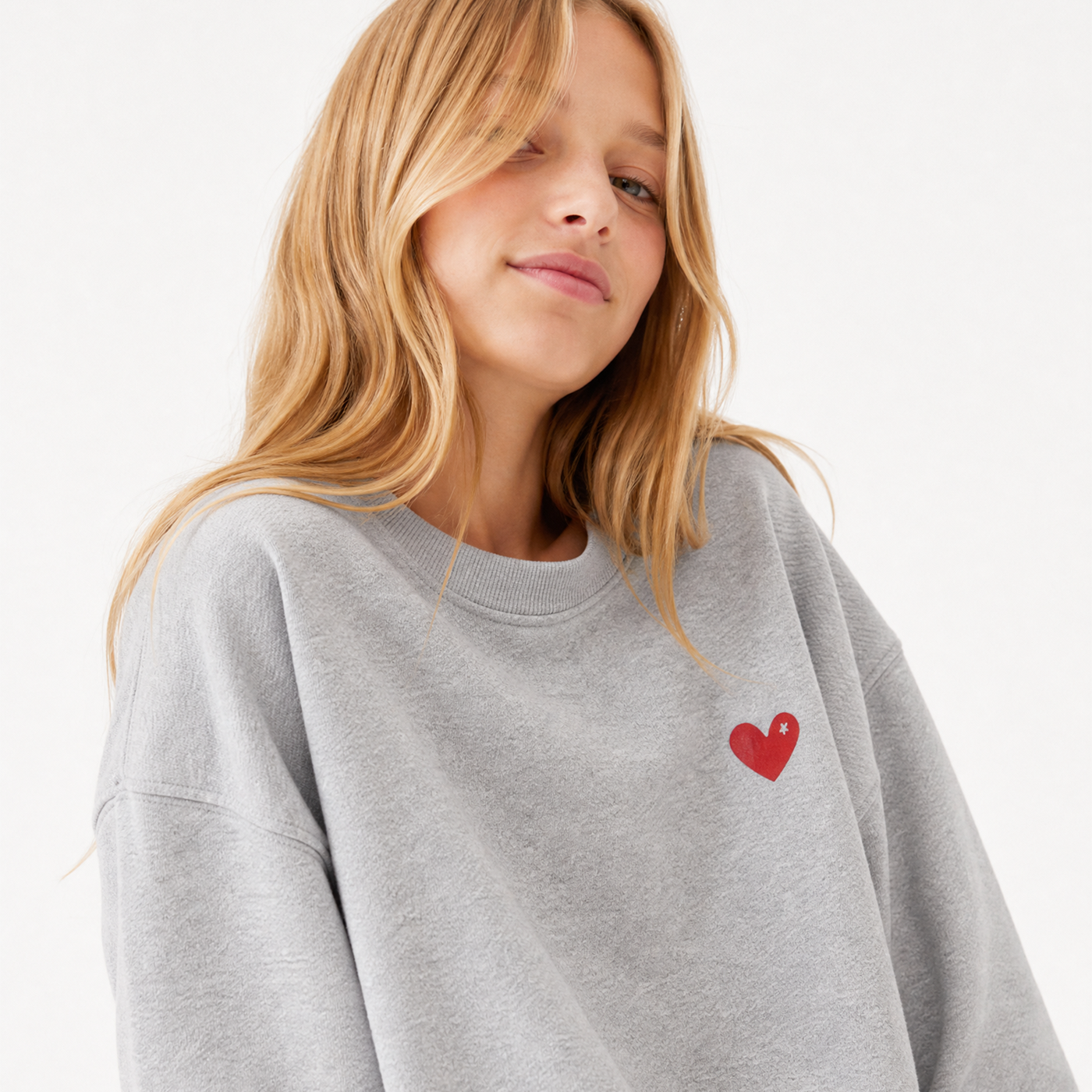 A woman wearing a light gray crewneck sweatshirt with a small red heart on the left chest, photographed against a clean white background.