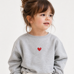 A young child wearing a light gray crewneck sweatshirt with a small red heart embroidered on the chest, photographed against a clean white background.
