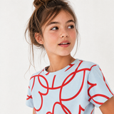 A young girl with her hair in a messy bun wearing a light blue t-shirt with a red abstract line pattern, photographed against a clean white background.