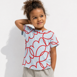 A young child wearing a light blue short-sleeve t-shirt with a red abstract line pattern, paired with striped pants, standing against a white wall.