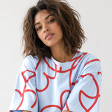 A woman with short, wavy brown hair wearing a light blue oversized t-shirt with a red abstract line pattern, photographed against a plain light gray background.